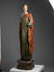 A tall, weathered wooden statue of a robed figure with a serene expression, draped in green and red garments, standing upon an octagonal base.