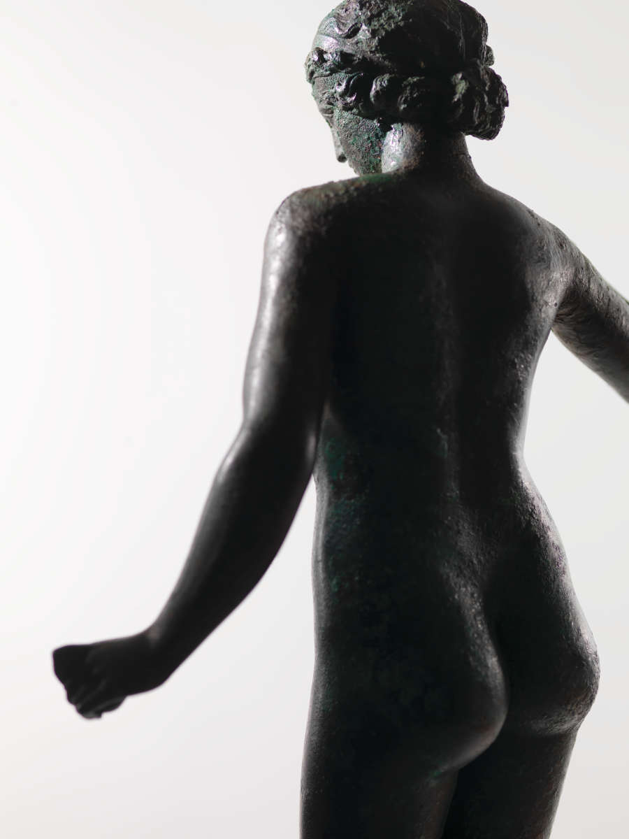 Back view of a tarnished bronze statue of a standing nude woman, her arms away from her body. The lighting amplifies the texture of the statue’s surface.