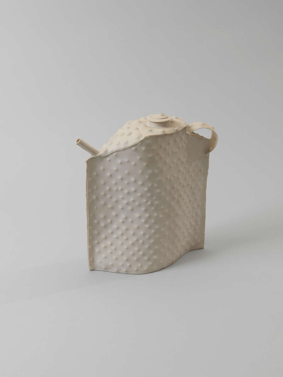 Minimalistic teapot with a dimpled surface, slightly flattened form, narrow spout, small curved handle, and a small coiled lid, all made in smooth, cream-colored matte ceramic.