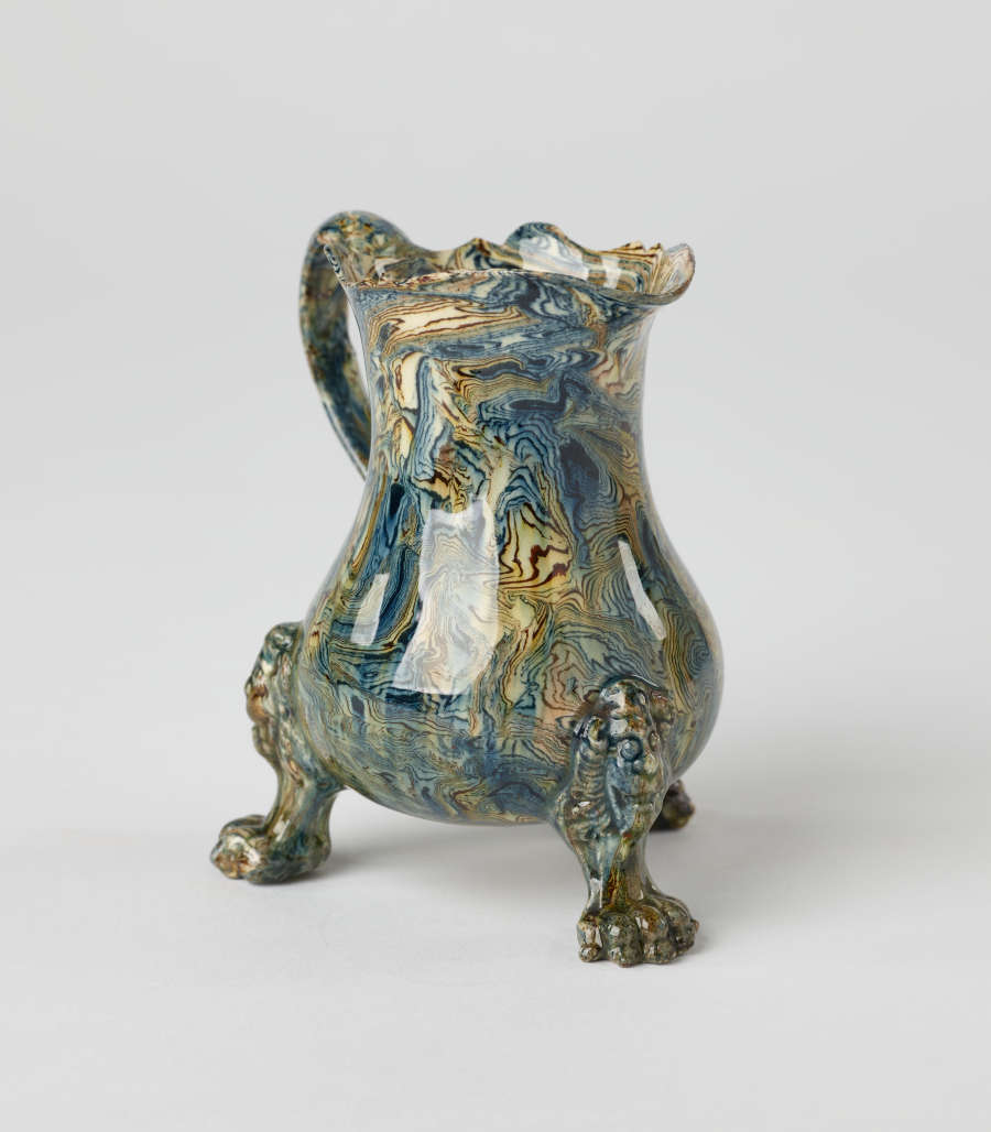 A small vessel with a cream, blue, and green mottled design. Three sculptural feet are shaped like lion’s paws with lion heads attached to body of vessel.