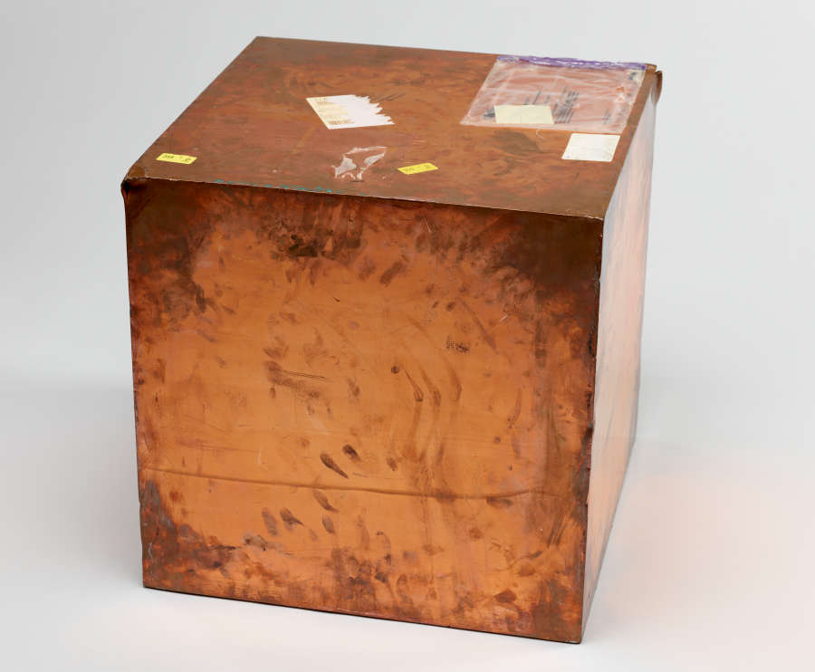 Image of Walead Beshty’s work: Copper box covered in various markings and shipping label stickers. The box is discolored in some areas.