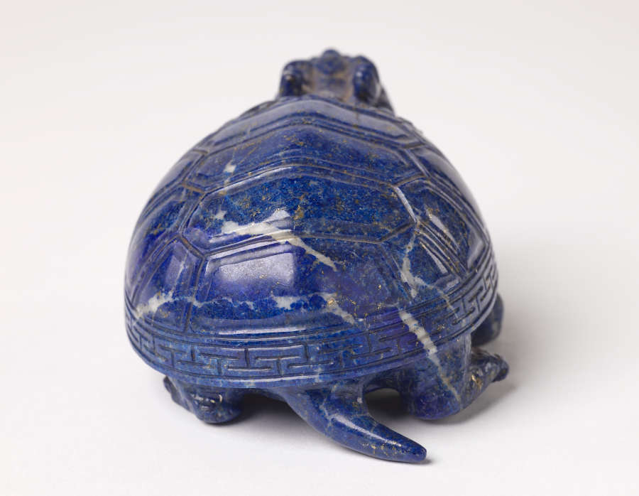 A back view of a blue sculpture of a horned tortoise, whose surface is covered with carved geometric patterns.