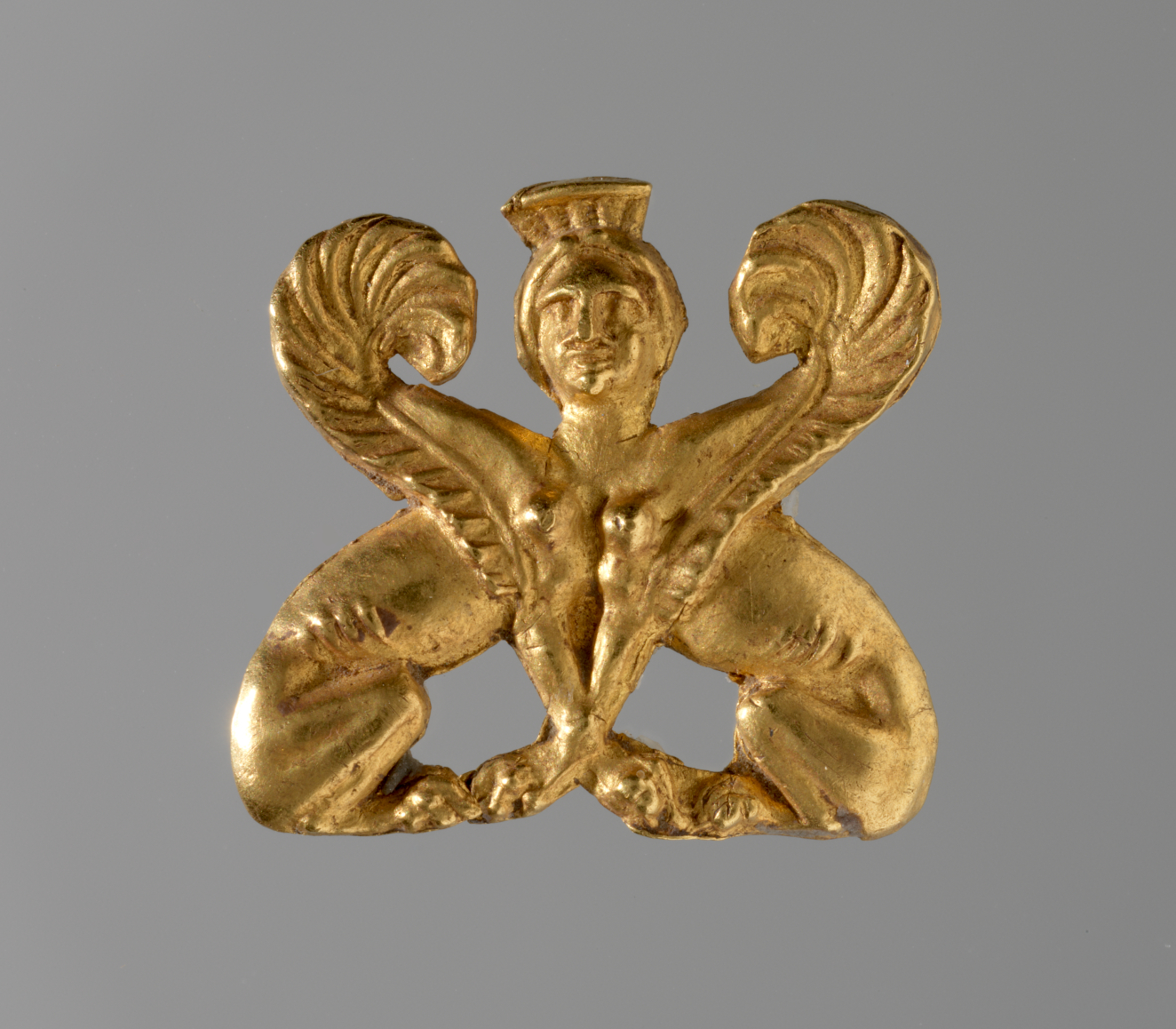 Gold ornament of two sphinx bodies facing one another. At their intersection is the gold crowned head of a woman, framed by two symmetrically arranged engraved wings.