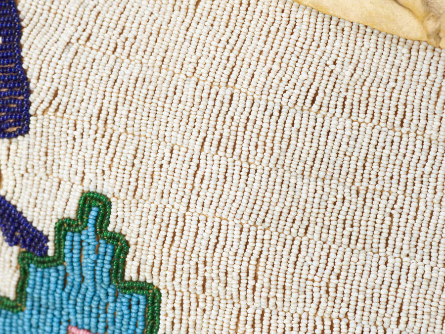 Detail of rows of white beads and a geometric blue decoration at bottom left. A small area of the cream leather ground shows at top. 
