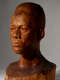 Three quarter view of a dramatically lit, glossy wooden bust depicting the head of a male figure with afrocentric facial features. He has a stern expression and an intense gaze.