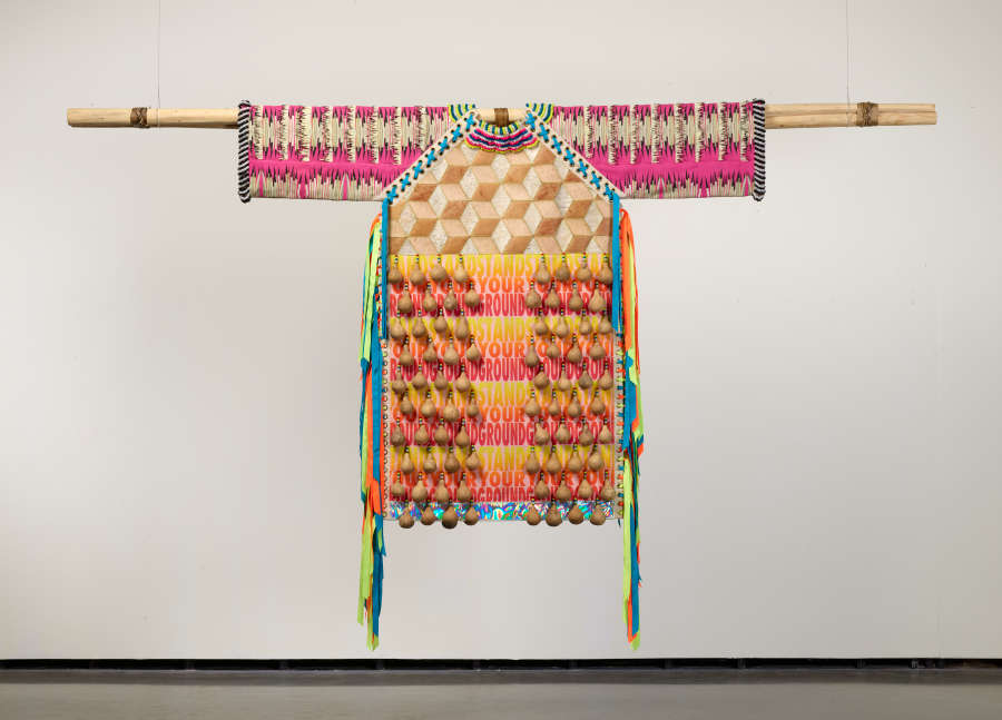 Multi-patterned, brightly colored, and tasseled shirt hanging on a wooden stick against a white background with machinery attached to its bottom. The floor is also shown.