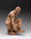 Realistically carved terracotta sculpture of a muscular man sitting on a rough pedestal and leaning over a vessel he’s holding between his legs which are spread to accommodate it.