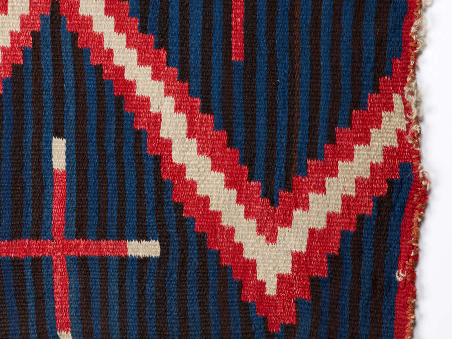 Closeup of a vertically striped black and blue woven blanket with a red-bordered, white zigzag pattern overtop and a frayed red trim. 