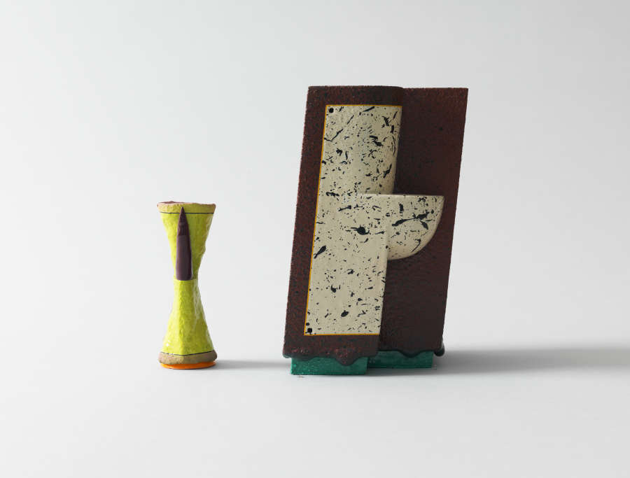 Two ceramic forms: a small yellow hourglass-shaped vessel with orange rim and dark central band beside a tilted rust red block featuring a cream speckled panel on a green base.