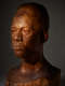 Three quarter view of a textured wooden bust depicting the head of a male figure with afrocentric facial features. He has a stern expression highlighted by high contrast lighting.
