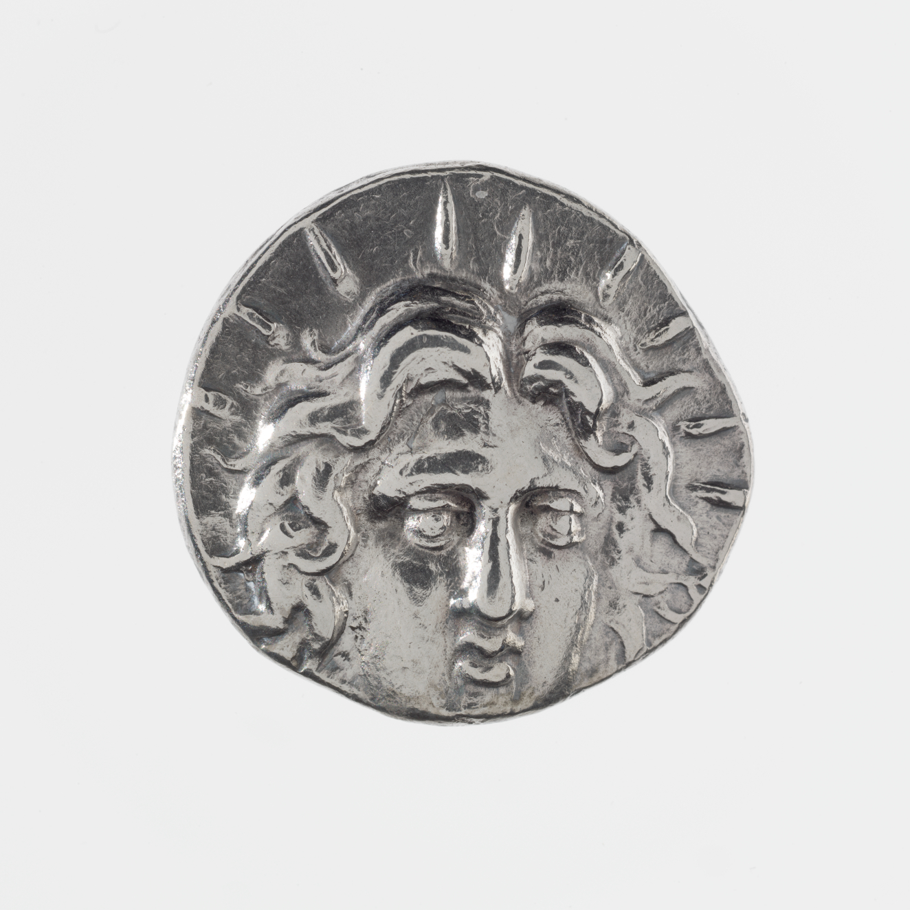 Silver coin with irregular edges. Embossed on its surface is a portrait of a person with wavy hair, lines emanating out from their profile.