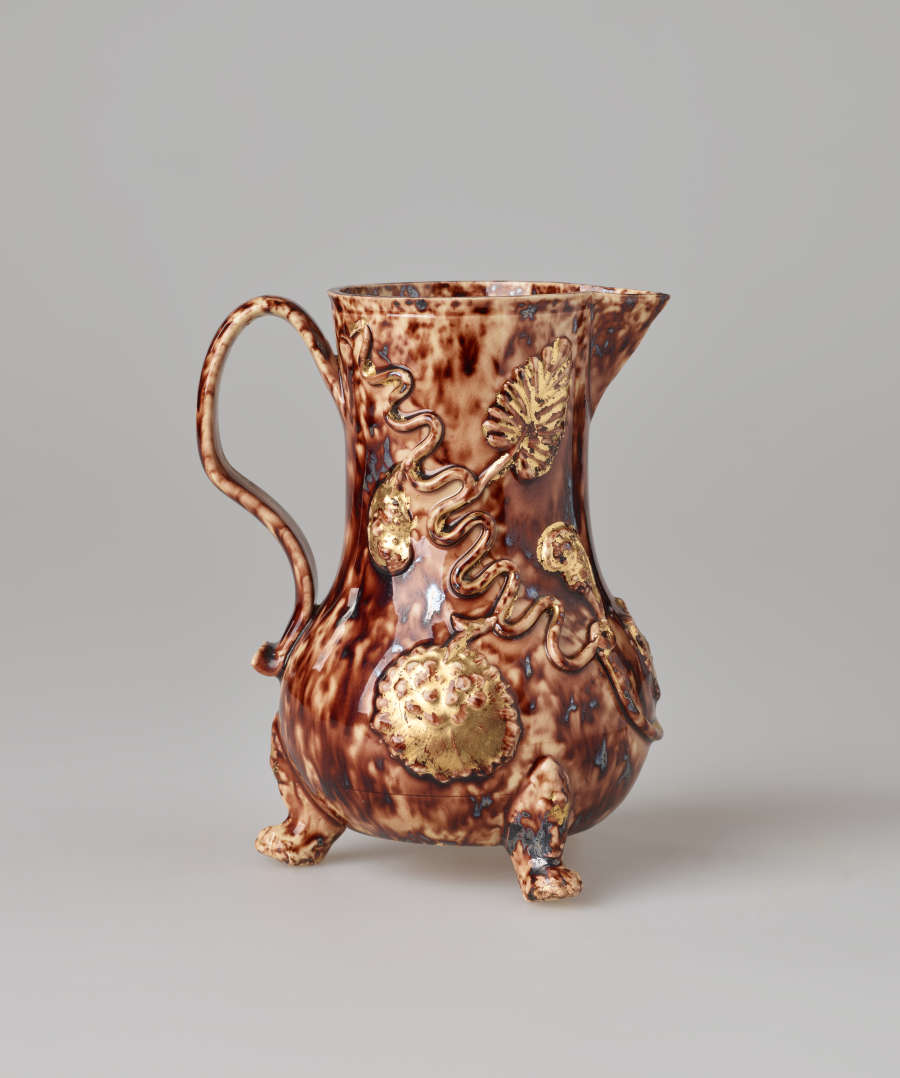 A mottled brown pitcher with a spout, handle, and multiple delicate feet. There are swirly gold floral decorations.
