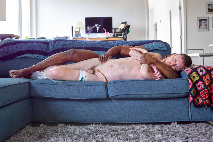 Color photograph of Coombs lying nude on a couch with his catheter visible above his penis. A Black man, whose face is covered by Coombs’s own, holds Coombs from behind.