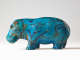 Side view of a bright turquoise hippopotamus decorated with black linework depicting reeds and papyrus fronds, its surface marked by splotches and lines showing the clay underneath the turquoise glaze.