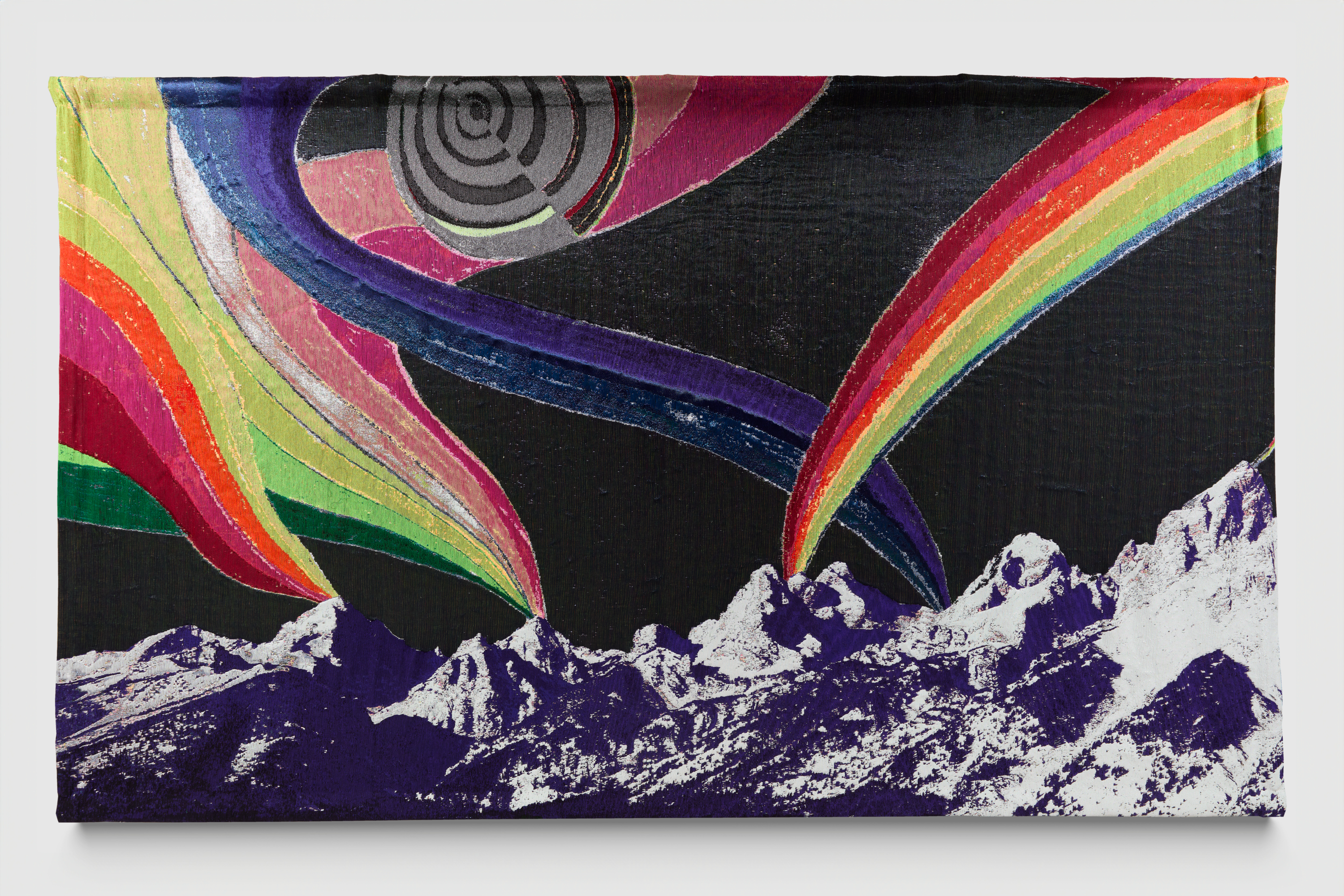 A woven wall-hanging with purple and white mountains in the foreground and vivid rainbows swirling out of the mountain peaks against a black sky. 
