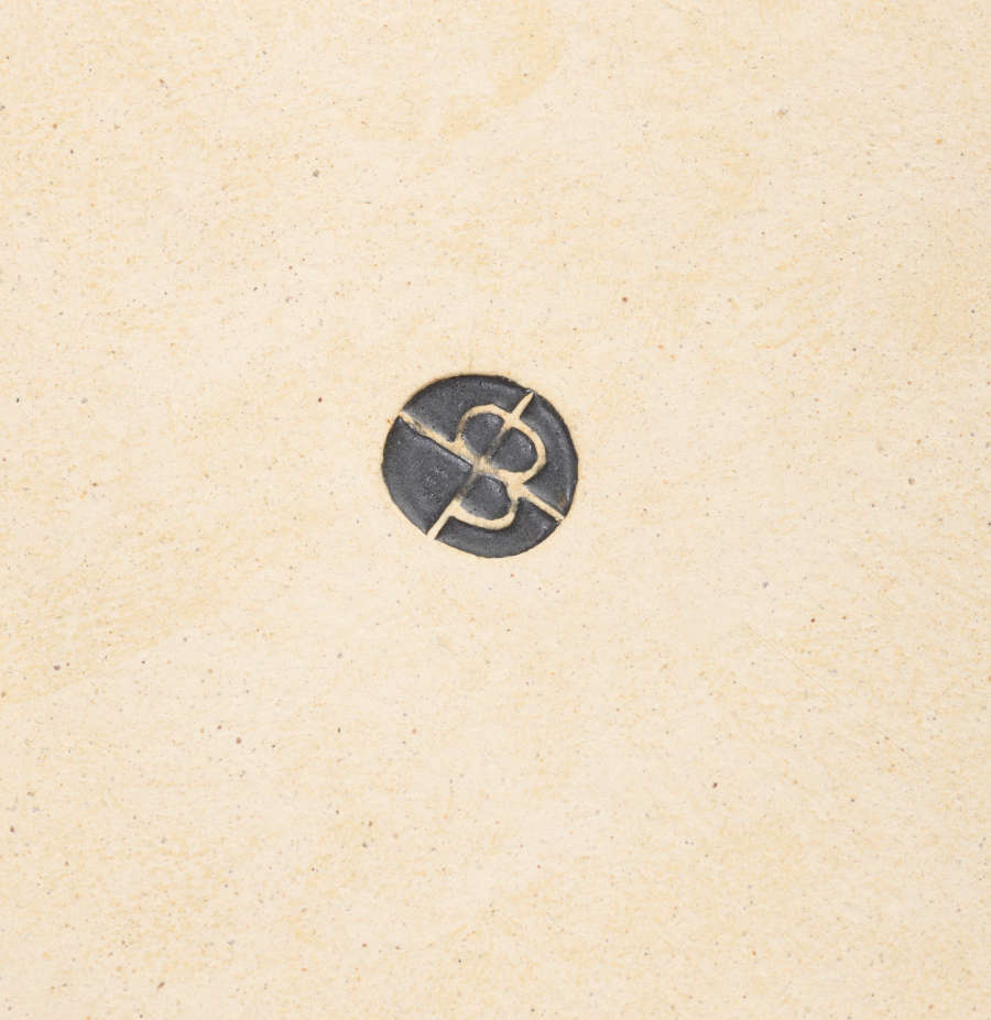 A small imprint on a beige ceramic surface. It features a decal that resembles an overlaid and reflected “P” and “B”, enclosed in a circle and filled with black glaze.