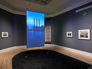 Hanging in the center of a gallery with dark-gray walls is a screen. The projected image shows an industrial building with three smokestacks. Artwork hangs on the surrounding walls.