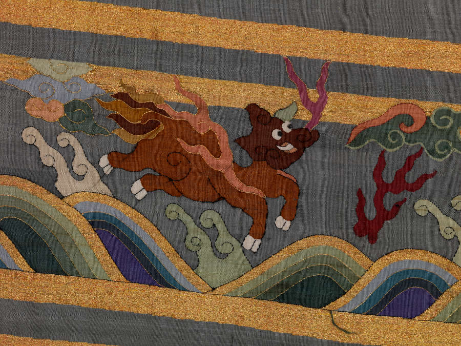 Border detail of the blue-gray robe’s back featuring a red animal leaping over waves, wispy pastel clouds and vegetal motifs encased in gold and blue stripe borders.