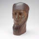 Three quarter front view of a dull wooden sculpture depicting a serene face wearing a draped cloth around its head. It features smooth contours and a vertical crack alongside its nose.