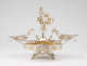 Elaborate silver double-oval-tray centerpiece with central winged-figurine holding up a gold garland, surrounded by cherubs, and framed by swirling arms extending to two shallow dishes, all elevated on four legs.