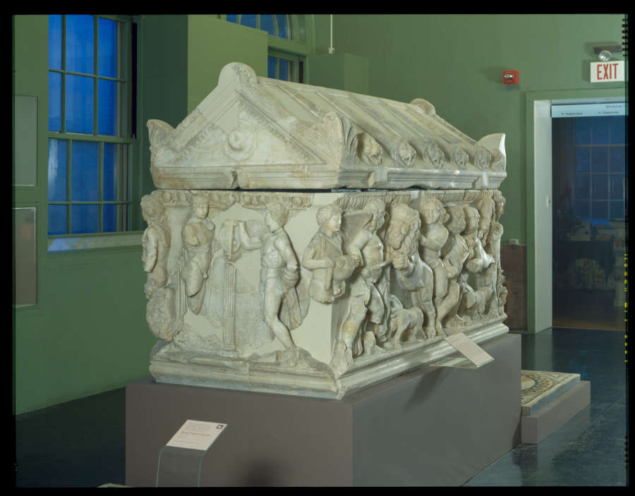 View of the whole sarcophagus in a gallery. The rectangular sides feature carvings of the Trojan war. The triangular lid is adorned with carved lion heads and floral details.