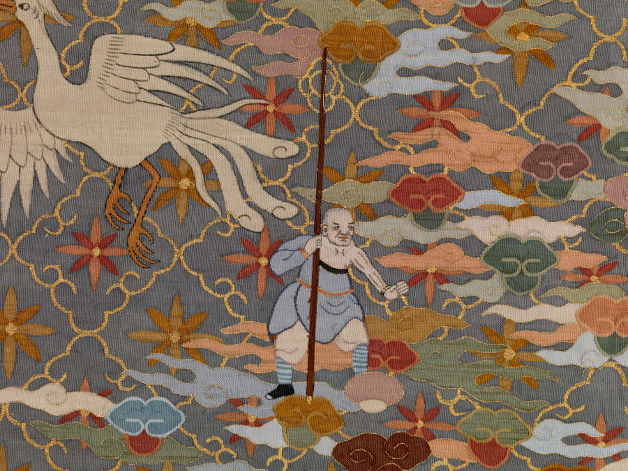 Robe’s back detail. A standing monk holds a tall staff amongst earthy pastel clouds and a soaring bird, against a blue-gray background with a diagonally running grid and floral motifs.
