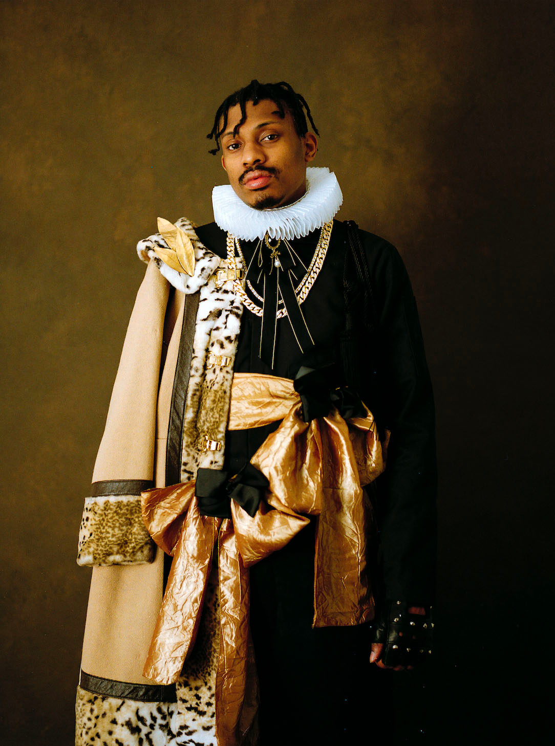 A photographic portrait of a dignified, dark-skinned figure staring out at the viewer, wearing a formal ensemble featuring a stiff collar, a fur-trimmed coat on one shoulder, two large, fabric bows, and a gold chain.