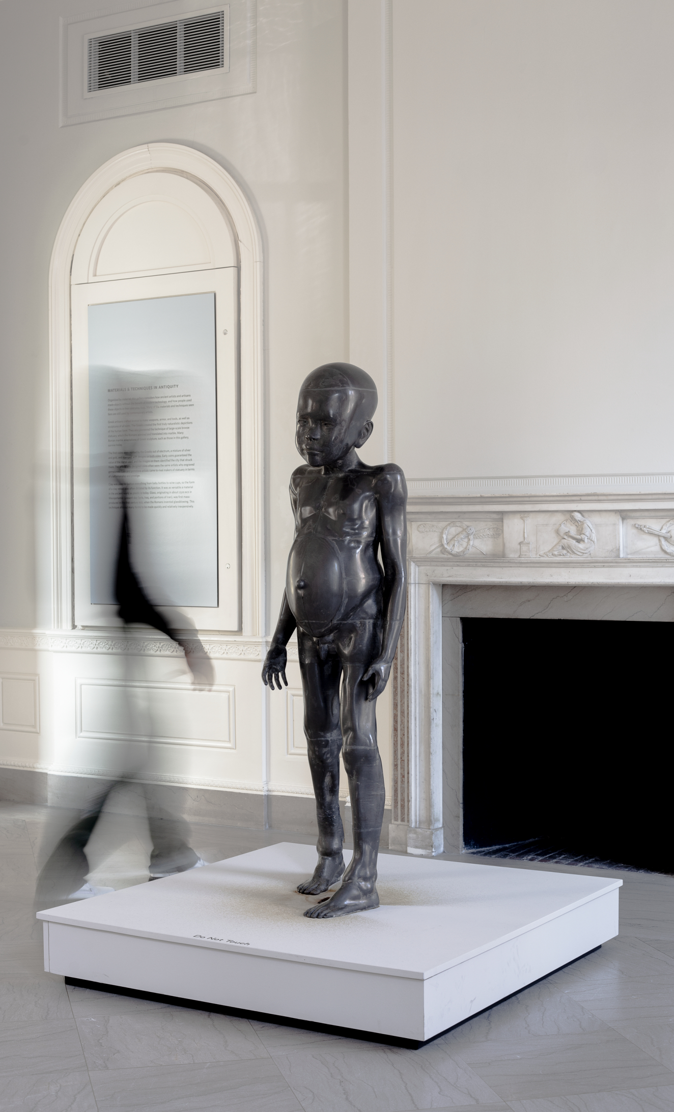 A vertical photograph of a sculpture of a naked, dark-skinned, child with a distended belly, swollen arms, and a bald head on a white, square platform in a museum gallery. There is the blurred person in motion walking on the left side of the platform. 