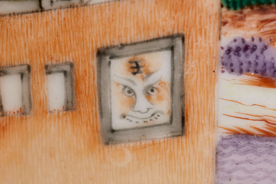 A close up of a painted ceramic depicting an orange wall with a window with an illustrated face, surrounded by smaller plain rectangular windows.