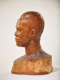 Profile view of a textured, but glossy, wooden-carved bust depicting a male figure with afrocentric facial features. The base is blurred, highlighting the details in the face and head.