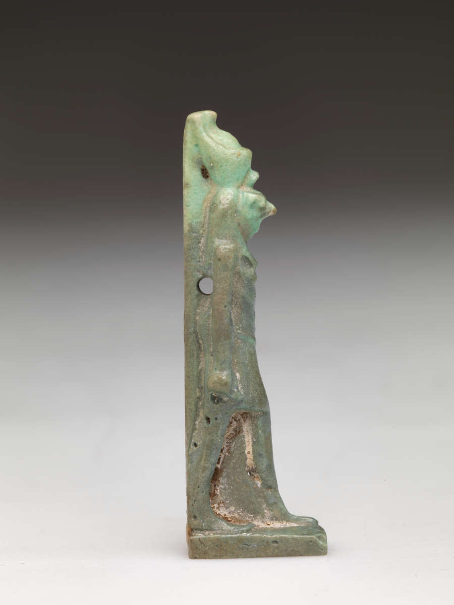 Side-view of a light-jade sculpture of a standing person with a tall head, narrow torso, and straight arms. The left leg has brown specks while the headdress is pale.