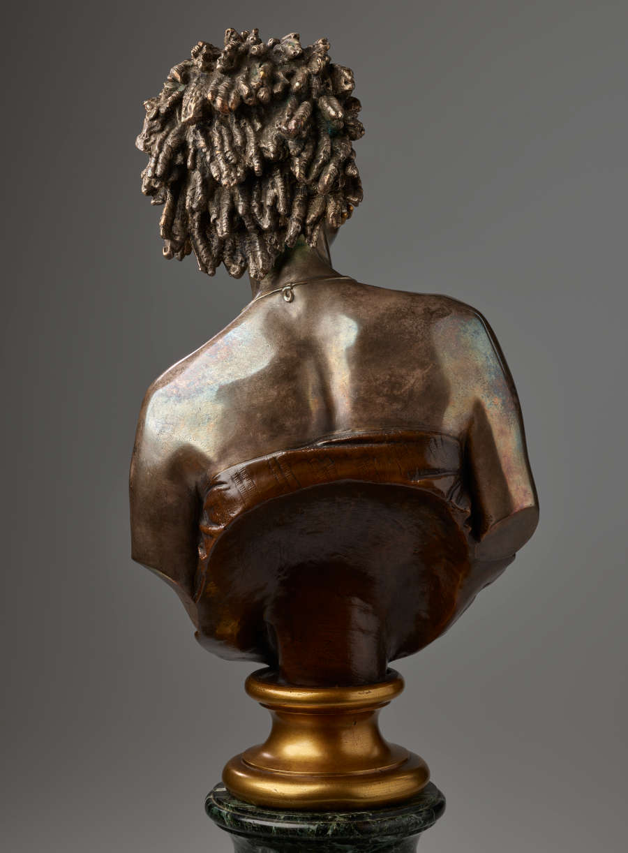Back view of a bronze bust highlighting hair in twists and sculpted shoulder blades exposed above a dark strapless top cut off diagonally and mounted on a gold base.