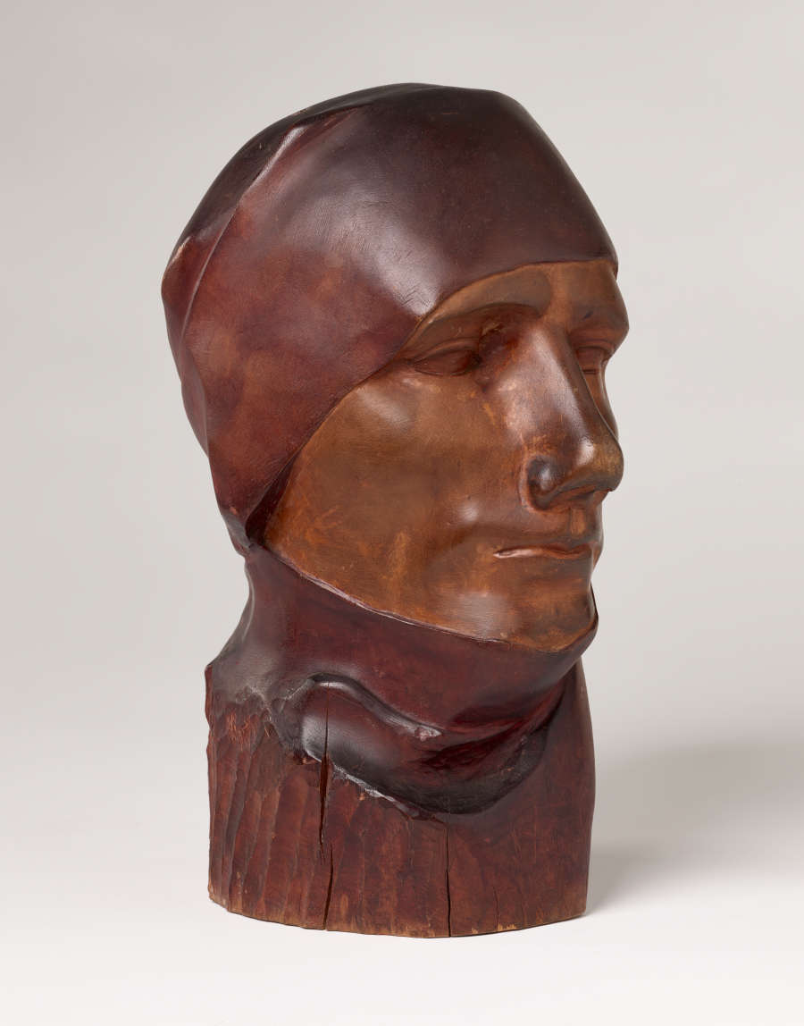 Three quarter front view of a vibrant, carved wood sculpture depicting a serene face wearing a cloth around its head and neck. The bust features smooth contours and a polished finish.