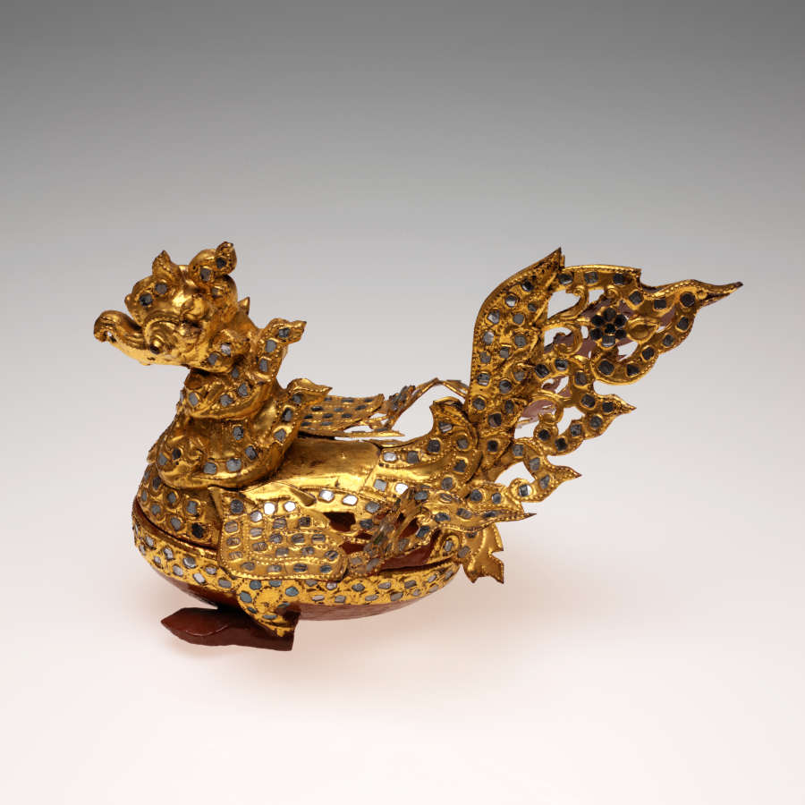 Opposite side-view of the golden, dragon-like, bird-shaped container. Its lid has a layered neck and body that shape upward, and its base is a cup-like stand supported by red feet.