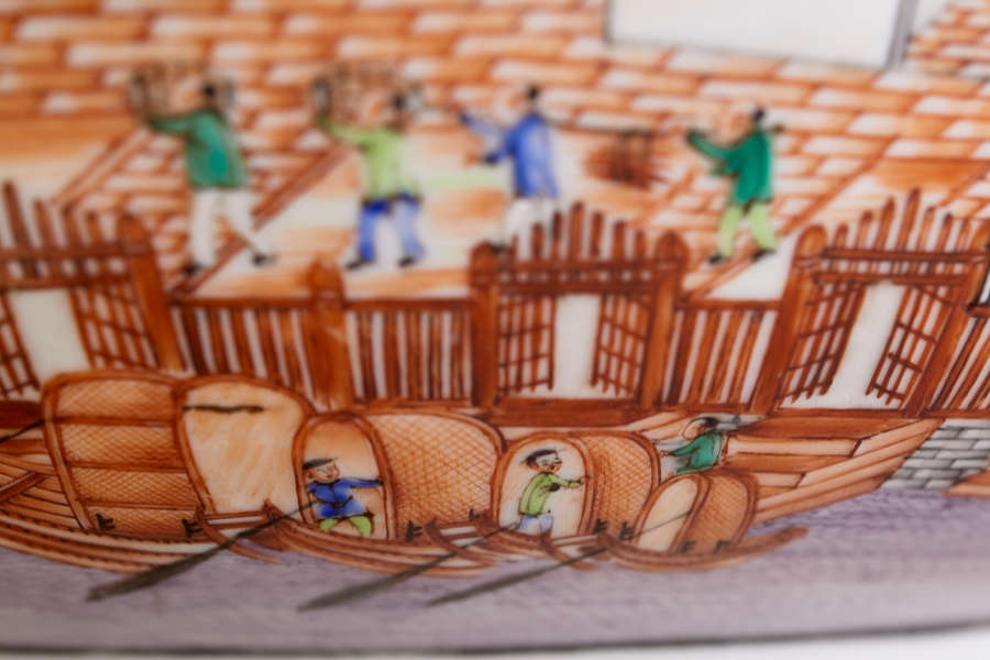 A close up of a painted ceramic depicting docked wooden boats in the foreground and workers carrying goods on an orange-tiled dock.