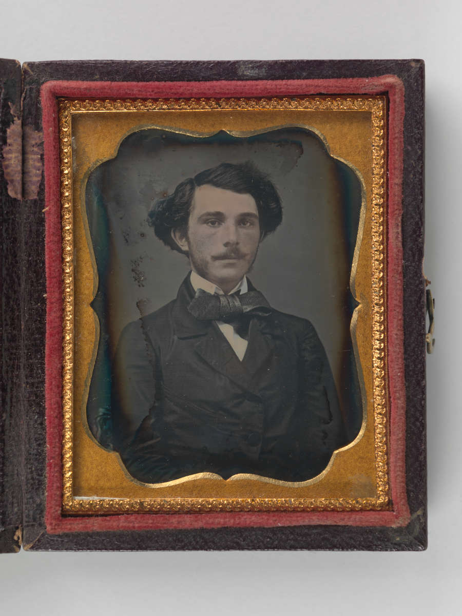 Vintage black and white portrait of a straight faced man in a suit and a bowtie, set in an ornate orange frame in a red and black box.