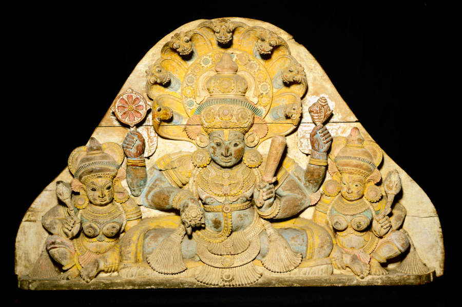 Rounded, cream-colored, triangular carving of three seated figures wearing ornate clothing and headdresses. The central figure is the largest and has four arms, each holding a different object.  
