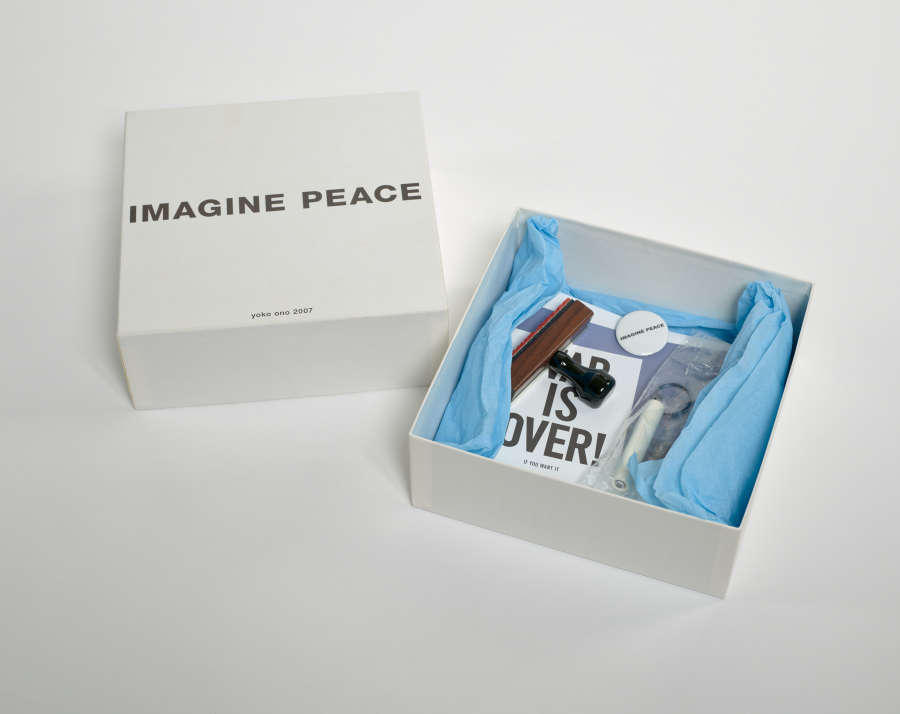 Open white square box. The box’s base is filled with various objects that are wrapped in blue tissue paper. The box top has bold black text that reads “Imagine Peace.”