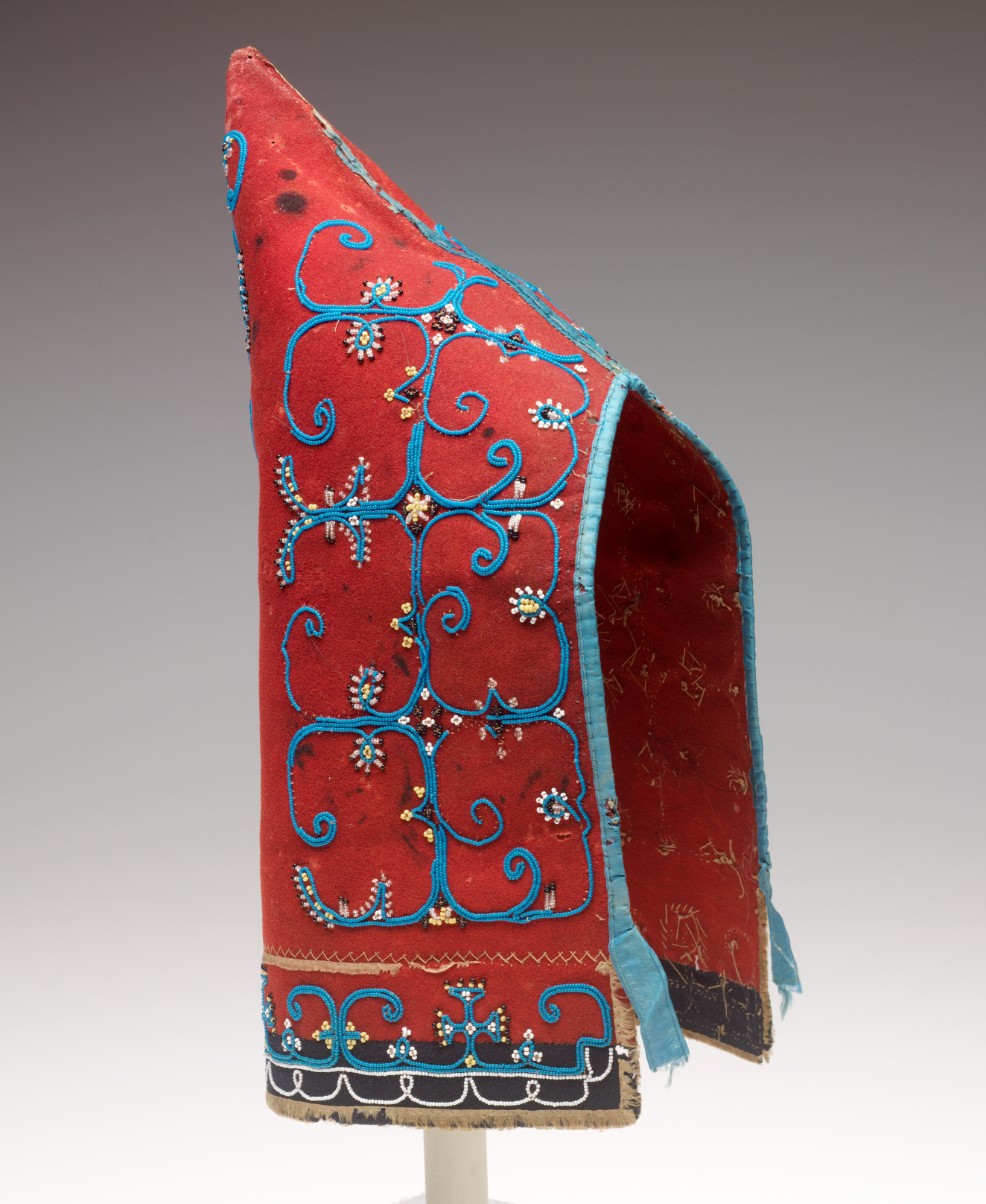 A tall, pointed conical red fabric hat with open front, featuring swirling blue embroidering, small white beads, and a slightly frayed blue-trimmed edge. 