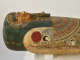Side view of a coffin with a gilded face, blue-black headdress, inlaid eyes, and a sculpted beard. A red sun above a black and white falcon sits on a shoulder.