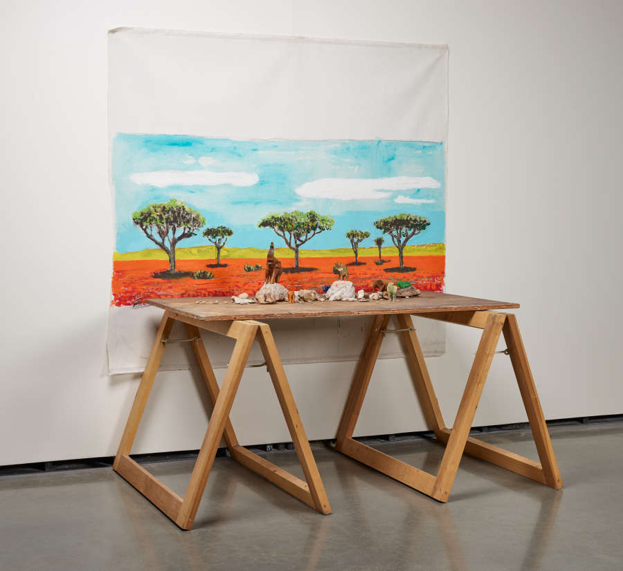 Side angle view of a white cloth painting of trees against a blue and orange background, hung behind a wooden table with objects on top.