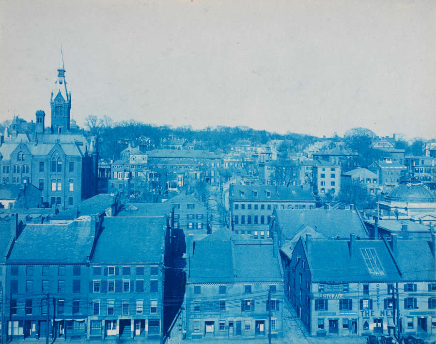 Blue-tinted vintage townscape, featuring a church with a prominent spire rising amidst dense streets of old brick buildings with gabled roofs and chimneys. The horizon is lined with shrubs.
