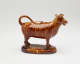  A brown sculptural creamer in the shape of a cow with horns. There is a small flat lid in the center of its back.