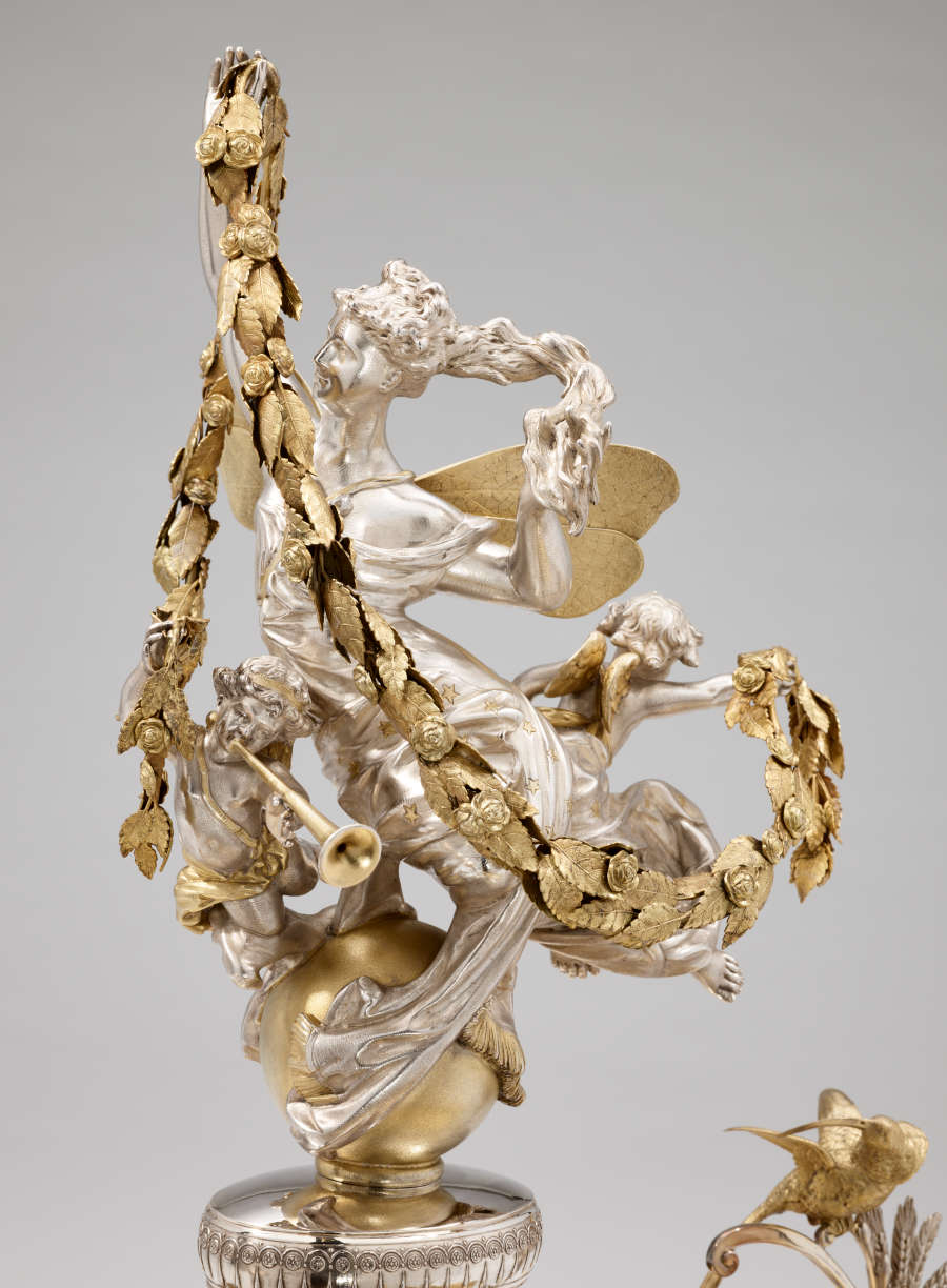 Silver-robed figure with flowing hair and gold wings holds a draping garland with one cherub on her back, holding the end of the garland while the other blows a horn.