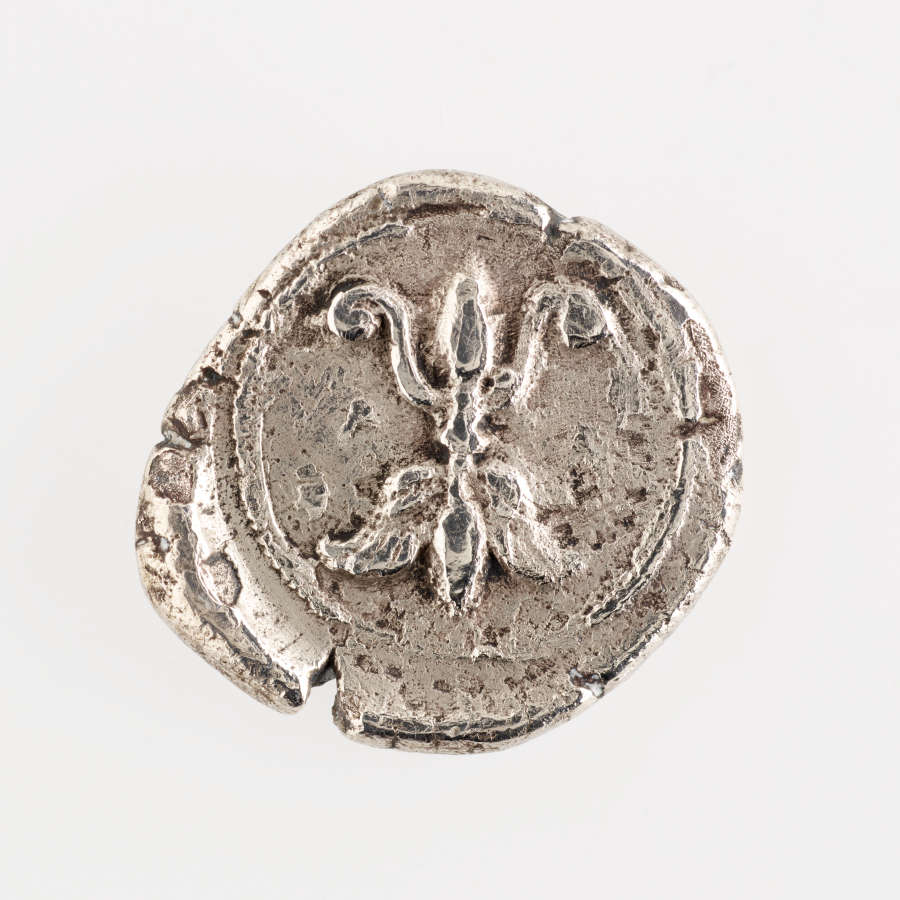 Shiny rough-edged, chipped, squarish silver coin with rounded corners, embossed with the fleur-de-lis motif, framed by a circular border.
