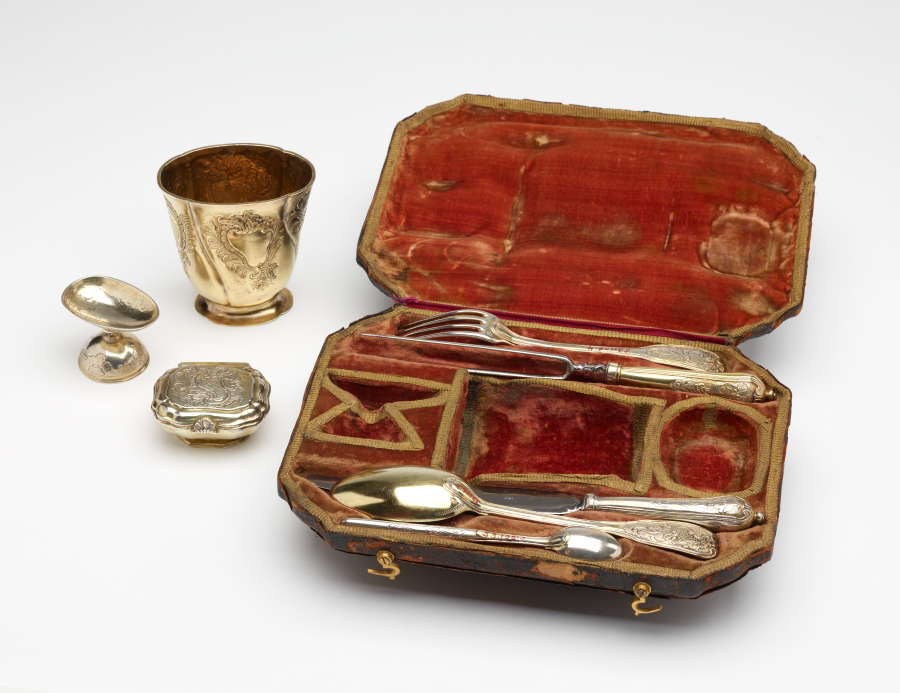 A traveling set with silverware. The traveling case has an orange colored fabric. There are multiple indents holding the silver, steel, and gilded cutlery and vessels.