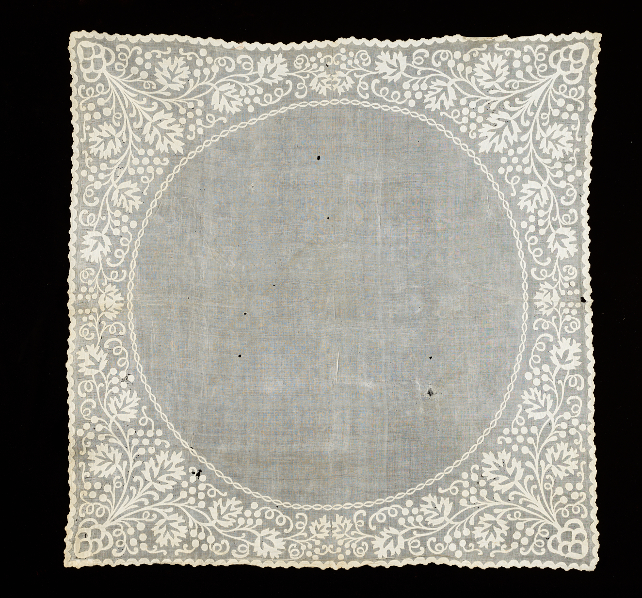 Light colored square fabric on a dark background. Features white embroidered floral patterns which point inwards from the corners and are worked around a large outlined circle of plain fabric.