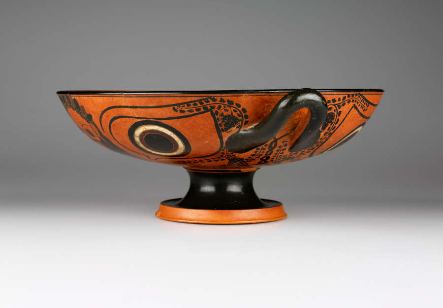Side view of a terracotta and black cup with a wide mouth, short black neck, and orange base. Its body features circular motifs rendered in black and white. 