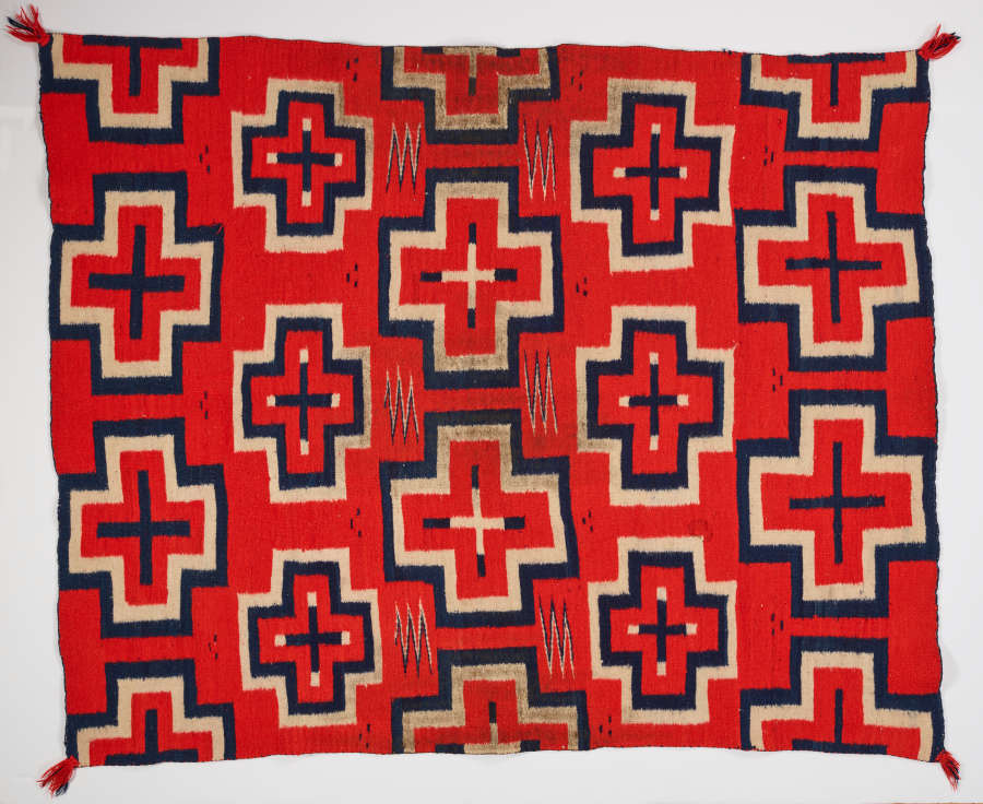 Red woven cloth with red-centered, geometric cross-shaped patterns throughout and red tassels in the corners. The navy blue and cream bordered crosses alternate between cream and navy blue.