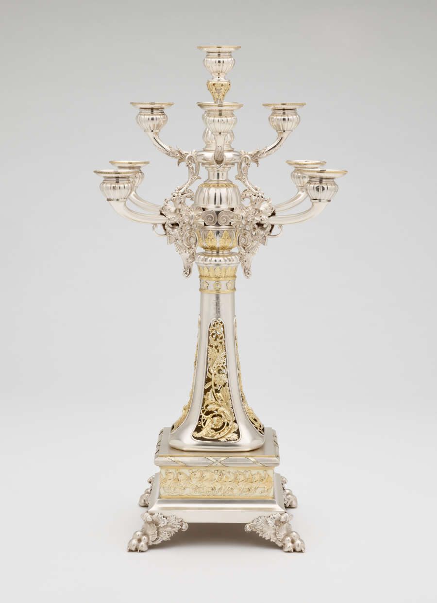 Front view of a silver nine branched candle holder, with detailed gold and ivory ornamentation of flowers and men on horseback.